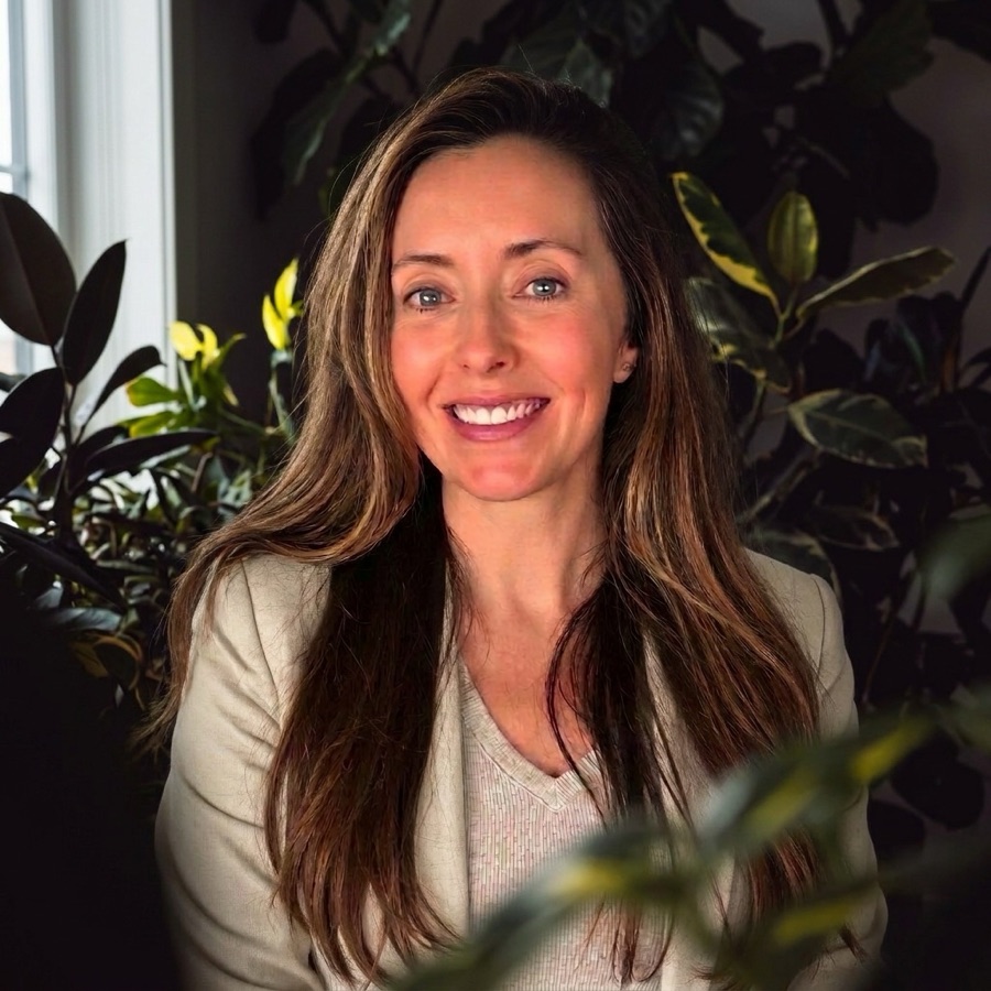 Sally Dehm, smiling, with long brown hair and a cream jacket, sitting among dark houseplants.